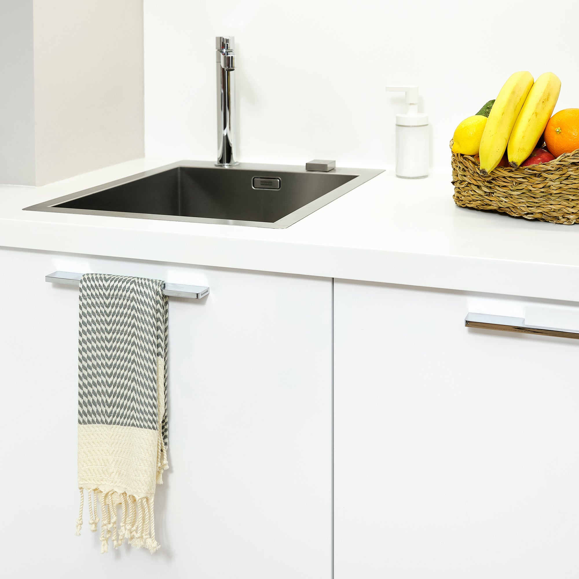 Modern kitchen with a sink, towel, and fruit basket on a white countertop.
