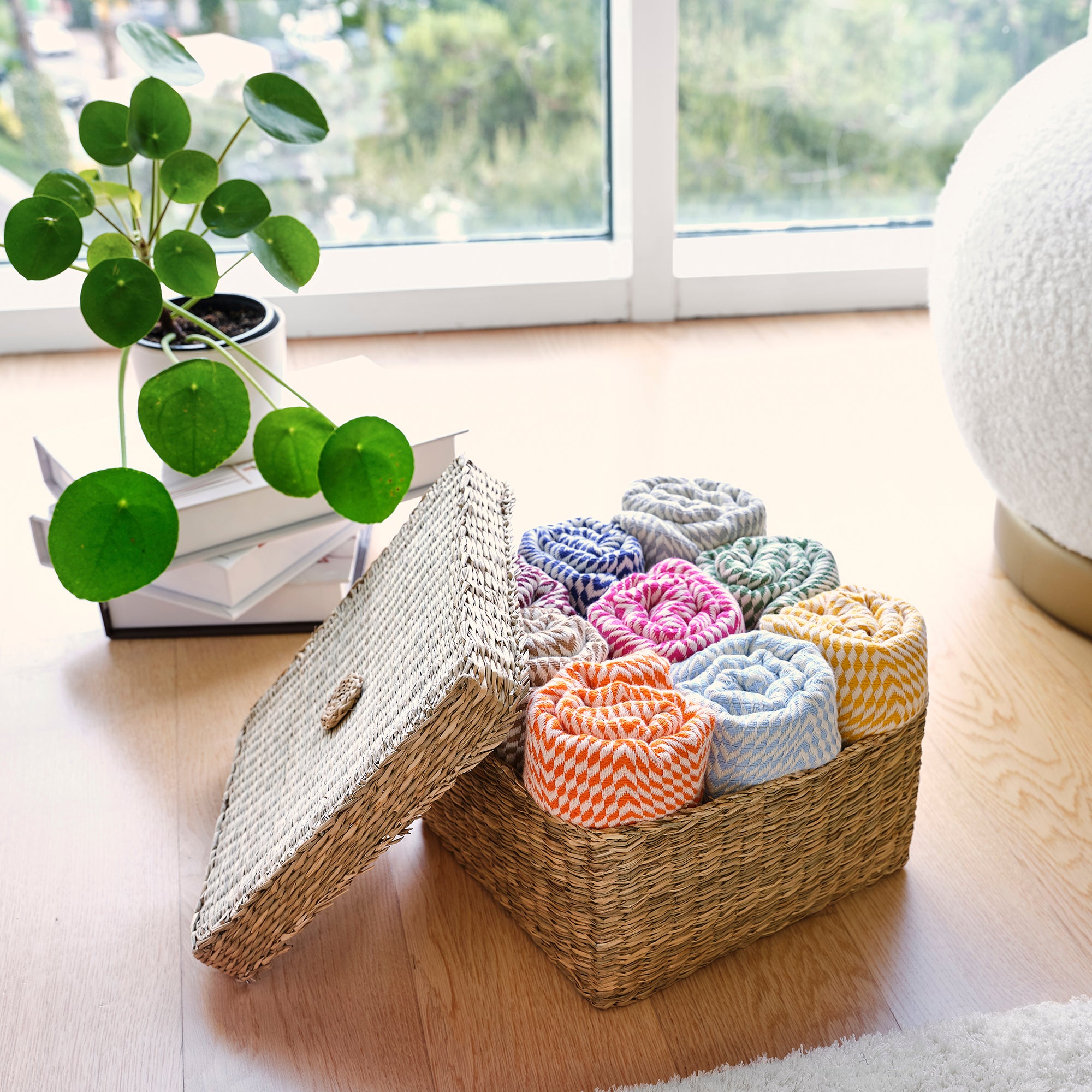 Wicker storage basket with colorful crochet items on a wooden floor next to a plant and books.
