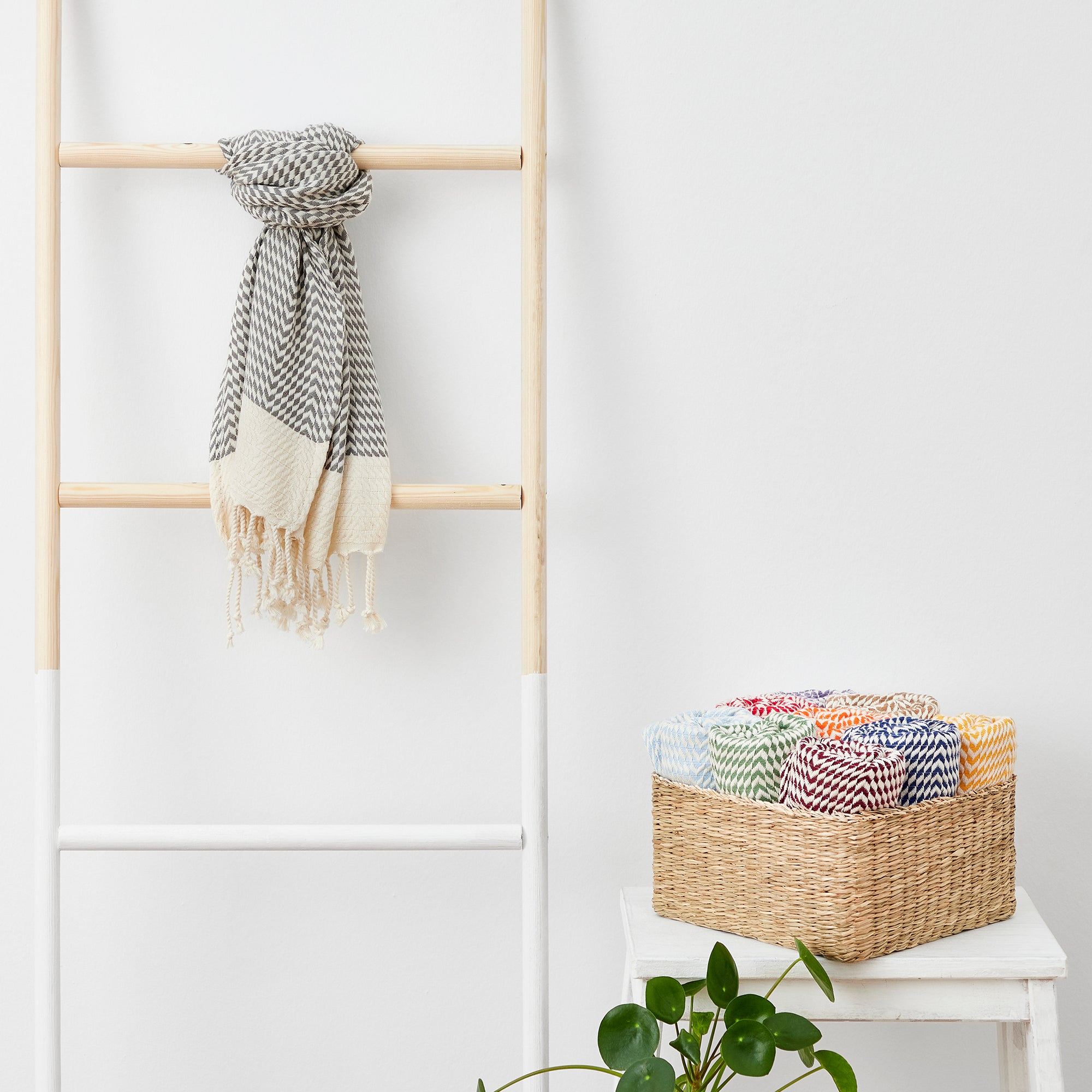 Scarf hanging on a wooden ladder with a basket of colorful jars below against a white wall.
